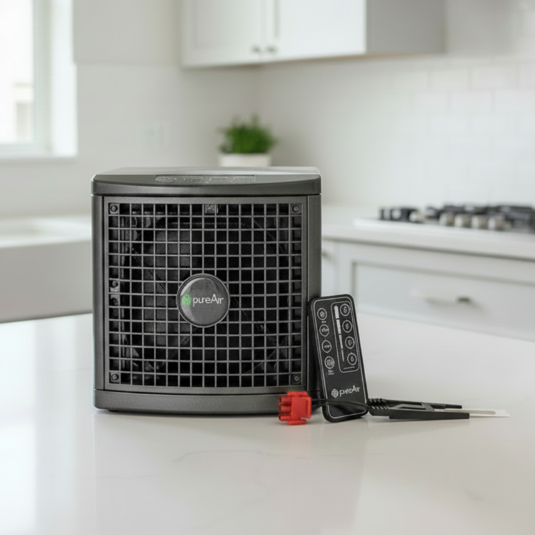 A compact PureAir 1500  dark gray sits on a white kitchen countertop with its remote control and replacement purification plate in front. The background shows a softly blurred modern kitchen interior.